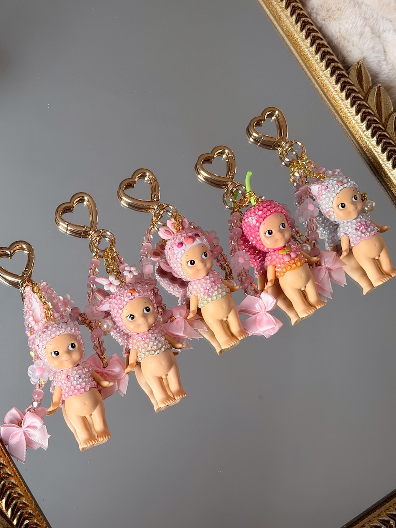 Puede incluir: Cinco llaveros con peque&ntilde;as figuras de mu&ntilde;ecas. Cada mu&ntilde;eca lleva un atuendo de cuentas rosas y blancas, alas y cierres dorados en forma de coraz&oacute;n. Los tocados var&iacute;an, incluyendo orejas de conejo y dise&ntilde;os florales. Cintas y cuentas rosas.