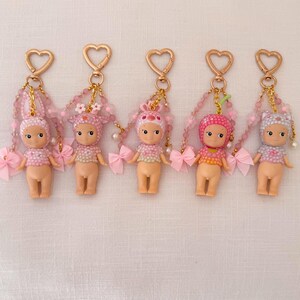 Puede incluir: Cinco peque&ntilde;os llaveros de mu&ntilde;ecas con cierres en forma de coraz&oacute;n. Cada mu&ntilde;eca tiene un atuendo y un tocado rosa o blanco diferentes. Cadenas de cuentas y lazos rosas cuelgan de los cierres.