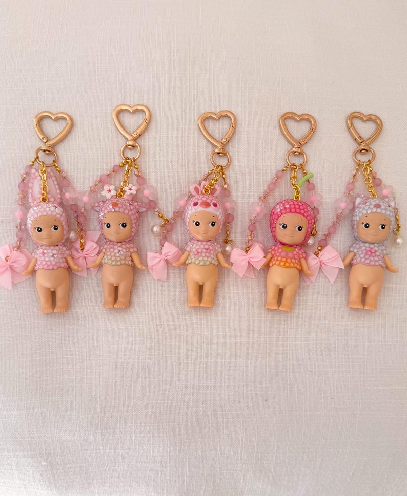 Puede incluir: Cinco peque&ntilde;os llaveros de mu&ntilde;ecas con cierres en forma de coraz&oacute;n. Cada mu&ntilde;eca tiene un atuendo y un tocado rosa o blanco diferentes. Cadenas de cuentas y lazos rosas cuelgan de los cierres.