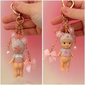 Puede incluir: Un llavero decorativo con una peque&ntilde;a mu&ntilde;eca con un atuendo de cuentas blanco y rosa y un sombrero a juego. La mu&ntilde;eca tiene un llavero dorado, un lazo rosa y detalles de perlas colgantes. El fondo es un degradado de rosa.