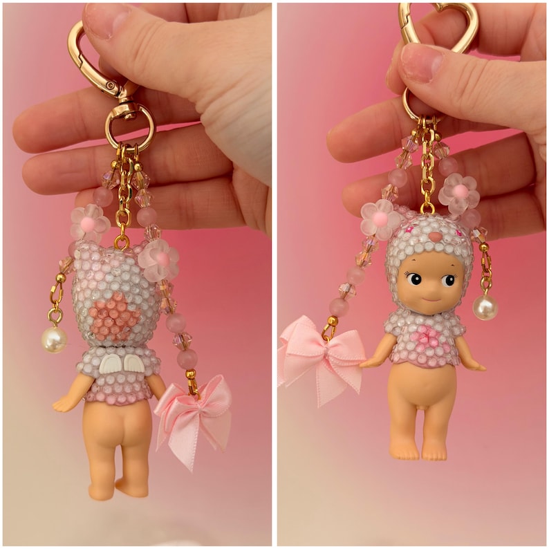 Puede incluir: Un llavero decorativo con una peque&ntilde;a mu&ntilde;eca con un atuendo de cuentas blanco y rosa y un sombrero a juego. La mu&ntilde;eca tiene un llavero dorado, un lazo rosa y detalles de perlas colgantes. El fondo es un degradado de rosa.