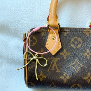 K&ouml;nnte beinhalten: Eine braune und beige Handtasche mit einem goldenen Schleifenanh&auml;nger, der mit einer rosa Schnur am Henkel befestigt ist.