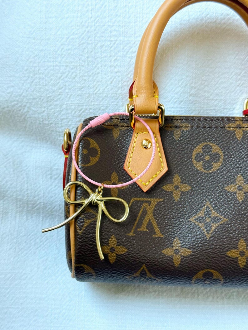 K&ouml;nnte beinhalten: Eine braune und beige Handtasche mit einem goldenen Schleifenanh&auml;nger, der mit einer rosa Schnur am Henkel befestigt ist.