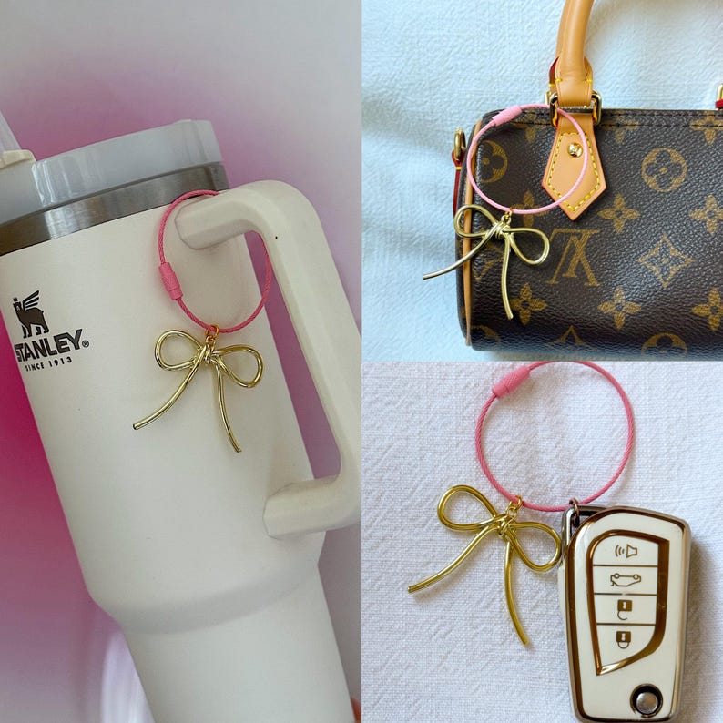 K&ouml;nnte beinhalten: Ein goldener Schleifenanh&auml;nger, der an einem rosafarbenen Gummiband befestigt ist. Der Anh&auml;nger ist an einer wei&szlig;en Stanley-Reiseflasche, einer braunen Ledertasche und einem wei&szlig;en Autoschl&uuml;ssel befestigt.