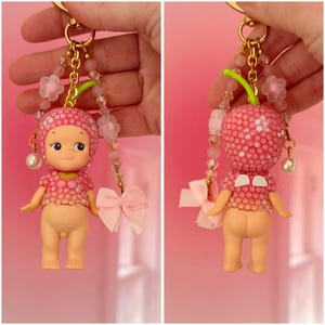 Puede incluir: Un llavero rosa con una figura de mu&ntilde;eca, cabeza en forma de cereza, cuentas rosas y un peque&ntilde;o lazo. La figura tiene una cara sonriente y est&aacute; unida a una cadena dorada con cuentas decorativas y un dije de perla.