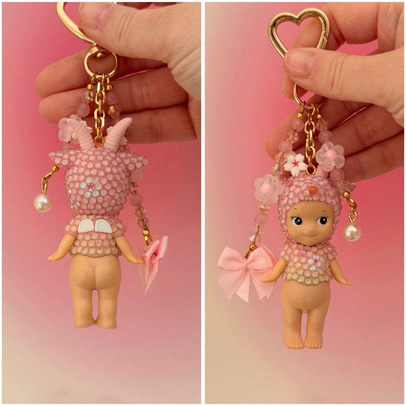 Puede incluir: Dos llaveros rosas con herrajes dorados. Uno presenta una figura de cabra y el otro una figura de beb&eacute;, ambos adornados con adornos rosas y perlas colgantes. Una cinta rosa est&aacute; unida a cada llavero.