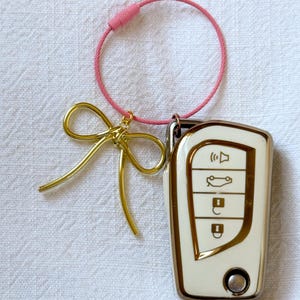 K&ouml;nnte beinhalten: Ein wei&szlig;er Autoschl&uuml;sselanh&auml;nger mit goldenen Akzenten und vier Kn&ouml;pfen. Der Schl&uuml;sselanh&auml;nger ist an einer rosa Schnur mit einer goldenen Schleife befestigt.