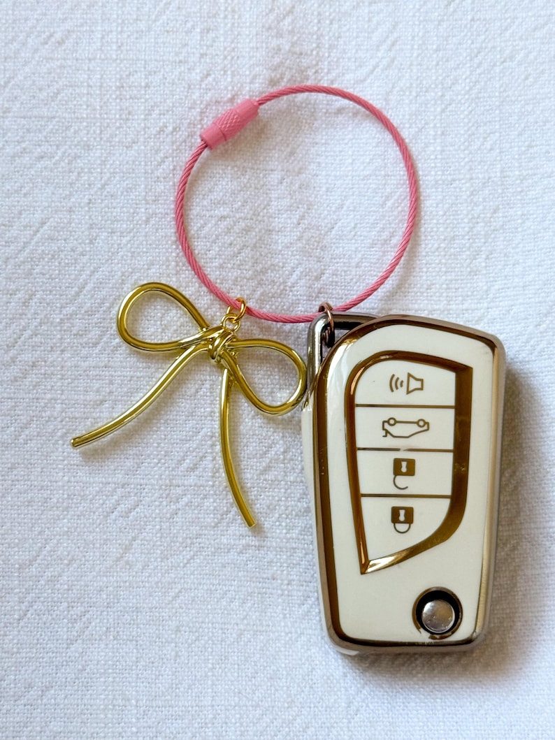 K&ouml;nnte beinhalten: Ein wei&szlig;er Autoschl&uuml;sselanh&auml;nger mit goldenen Akzenten und vier Kn&ouml;pfen. Der Schl&uuml;sselanh&auml;nger ist an einer rosa Schnur mit einer goldenen Schleife befestigt.