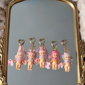 Puede incluir: Cinco peque&ntilde;os llaveros de mu&ntilde;ecas con detalles rosas y blancos se exhiben en un espejo con marco dorado. Cada mu&ntilde;eca tiene un sombrero &uacute;nico y detalles de cuentas, con lazos rosas y cierres en forma de coraz&oacute;n.