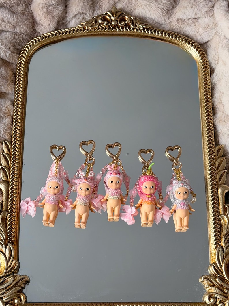 Puede incluir: Cinco peque&ntilde;os llaveros de mu&ntilde;ecas con detalles rosas y blancos se exhiben en un espejo con marco dorado. Cada mu&ntilde;eca tiene un sombrero &uacute;nico y detalles de cuentas, con lazos rosas y cierres en forma de coraz&oacute;n.