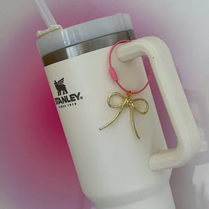 K&ouml;nnte beinhalten: Wei&szlig;e Stanley-Reisebecher mit einem rosa und goldenen Schleifenanh&auml;nger an der Henkel. Der Becher hat ein schwarzes Logo mit der Aufschrift "STANLEY" und "Since 1913".