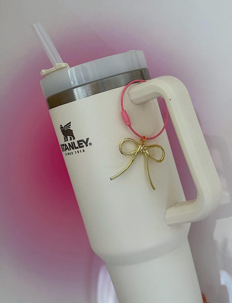 K&ouml;nnte beinhalten: Wei&szlig;e Stanley-Reisebecher mit einem rosa und goldenen Schleifenanh&auml;nger an der Henkel. Der Becher hat ein schwarzes Logo mit der Aufschrift "STANLEY" und "Since 1913".