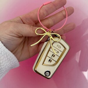 K&ouml;nnte beinhalten: Ein goldener Schl&uuml;sselanh&auml;nger mit einem wei&szlig;en Autoschl&uuml;ssel und einer goldenen Schleife. Der Schl&uuml;sselanh&auml;nger ist an einer rosa Schnur befestigt.