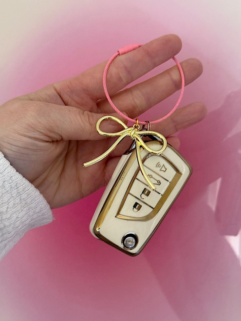 K&ouml;nnte beinhalten: Ein goldener Schl&uuml;sselanh&auml;nger mit einem wei&szlig;en Autoschl&uuml;ssel und einer goldenen Schleife. Der Schl&uuml;sselanh&auml;nger ist an einer rosa Schnur befestigt.