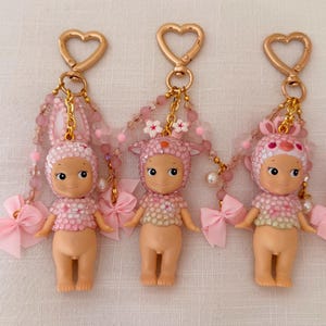 Puede incluir: Tres llaveros de mu&ntilde;ecas rosas con cierres dorados en forma de coraz&oacute;n. Cada mu&ntilde;eca tiene un sombrero con tem&aacute;tica de animal diferente y un lazo rosa. Los llaveros presentan detalles de cuentas rosas y se muestran sobre una superficie blanca.