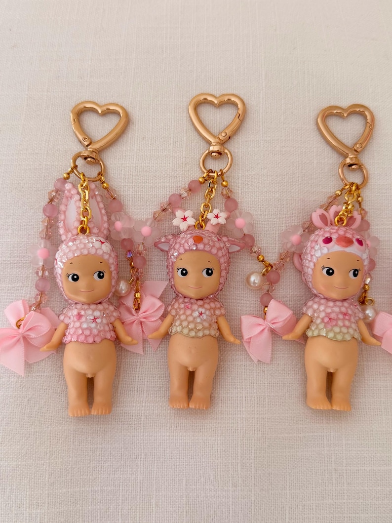 Puede incluir: Tres llaveros de mu&ntilde;ecas rosas con cierres dorados en forma de coraz&oacute;n. Cada mu&ntilde;eca tiene un sombrero con tem&aacute;tica de animal diferente y un lazo rosa. Los llaveros presentan detalles de cuentas rosas y se muestran sobre una superficie blanca.