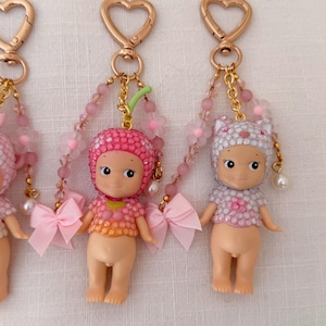 Puede incluir: Tres peque&ntilde;os llaveros con forma de mu&ntilde;eca, con cierres dorados en forma de coraz&oacute;n. Cada mu&ntilde;eca tiene un sombrero diferente, incluyendo un dise&ntilde;o de fresa y gato. Cuentan con detalles en rosa y blanco, cadenas de cuentas y lazos rosas.