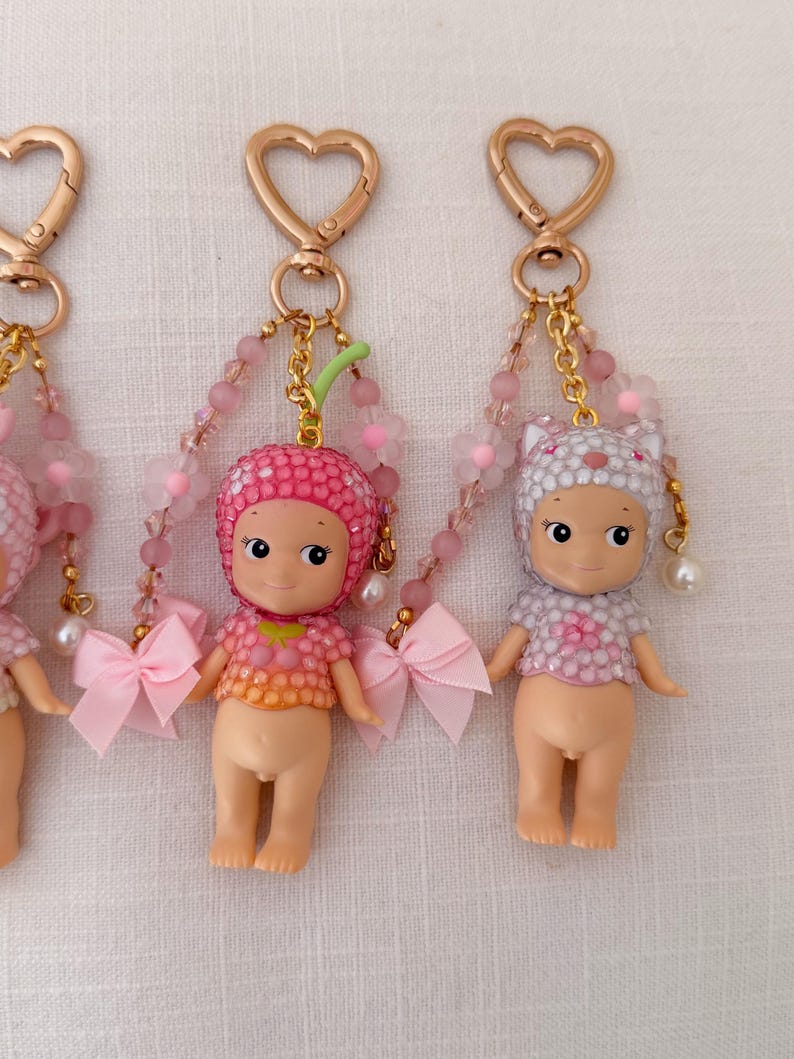 Puede incluir: Tres peque&ntilde;os llaveros con forma de mu&ntilde;eca, con cierres dorados en forma de coraz&oacute;n. Cada mu&ntilde;eca tiene un sombrero diferente, incluyendo un dise&ntilde;o de fresa y gato. Cuentan con detalles en rosa y blanco, cadenas de cuentas y lazos rosas.