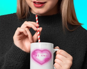 May include: White ceramic mug with a pink heart design and the text "BE KIND" in a neon pink font. The mug is being held by a person with a red and white striped straw in their hand.