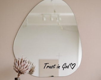 May include: An oval wall mirror with the text "Trust in God" and a heart symbol. A dried flower arrangement in a brown vase is in front of the mirror, set against a neutral-toned wall.