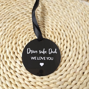May include: A black circular ornament with the text "Drive safe Dad WE LOVE YOU" in white, and a white heart symbol. It has a black ribbon for hanging. The ornament is on a woven surface.