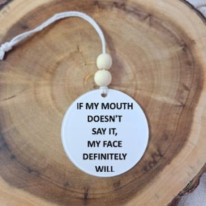 Peut inclure: Un ornement circulaire blanc portant le texte noir IF MY MOUTH DOESN'T SAY IT, MY FACE DEFINITELY WILL. L'ornement est fixé à une ficelle blanche avec deux petites perles en bois rondes et claires au-dessus du disque circulaire.