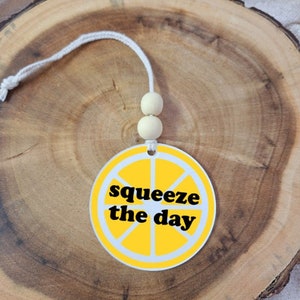 Peut inclure: Un désodorisant rond jaune et blanc avec un motif de tranche de citron et l'inscription "squeeze the day" en noir. Il est suspendu à une ficelle blanche avec deux perles en bois.