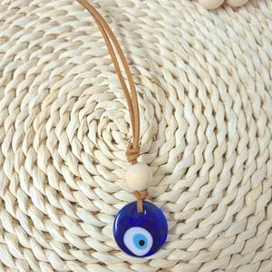 May include: A blue glass evil eye pendant necklace with a brown leather cord. The pendant has a white and black eye design. The necklace also features a wooden bead and is displayed on a woven straw background.