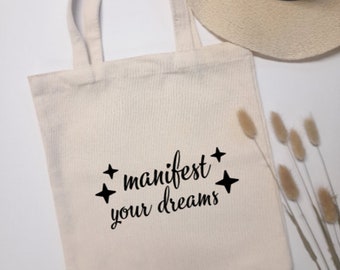 May include: A cream-coloured tote bag featuring the words "manifest your dreams" in black script, with four black star shapes. A straw hat and dried flowers are in the background.