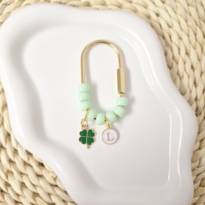 May include: A gold-toned U-shaped metal keyring featuring a series of light mint green cylindrical beads. Two small charms hang from the beads: a dark green four-leaf clover and a circular white charm with the letter L printed in gold.