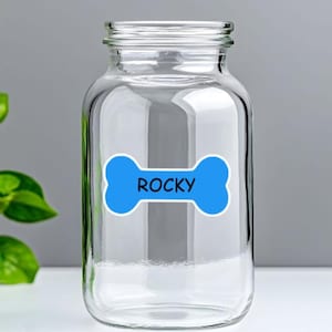 May include: A clear glass jar with a wide opening, displaying a blue bone-shaped sticker with the name "ROCKY" in white text. The jar is empty and rests on a white surface.