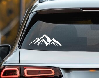 May include: White mountain range decal applied to the rear window of a silver car. The decal features a stylized outline of jagged mountain peaks, creating a minimalist outdoor theme. The car's taillights are visible below the window.