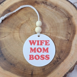 May include: A white, circular car air freshener with the words "WIFE MOM BOSS" in red block letters. It has a white string and two wooden beads for hanging. The background is a wooden surface.