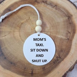 Peut inclure: Un ornement circulaire blanc portant le texte noir "MOM'S TAXI. SIT DOWN AND SHUT UP." L'ornement est suspendu par un cordon blanc orné de deux petites perles en bois de couleur claire, créant une décoration suspendue simple et fonctionnelle.
