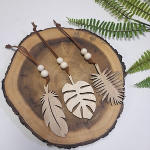 May include: Three wooden leaf and feather ornaments with leather cords and wooden beads. The ornaments are displayed on a round, wooden slice with a natural edge. Green fern-like leaves are in the background.