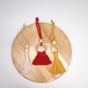 May include: Three macrame car air fresheners in cream, red, and yellow. Each has a wooden ring and bead, with a tassel. The air fresheners are displayed on a round, light-coloured wooden surface.