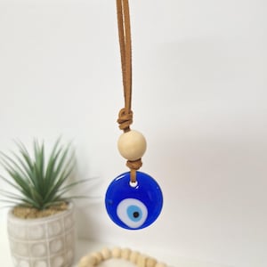 May include: A blue glass evil eye charm with a white and black eye design, hanging from a brown leather cord. A wooden bead sits above the charm. The charm is a decorative item.