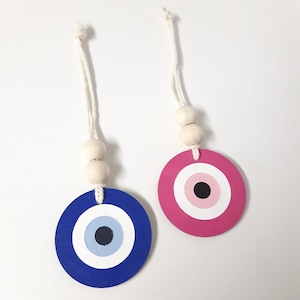 May include: Two circular eye-shaped ornaments. One is blue with a light blue and white centre, the other is pink with a pink and white centre. Each has a white string with wooden beads.