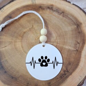 May include: A circular white ornament featuring a black paw print design within a heartbeat line. The paw print has a heart in the middle. It hangs from a white string with two wooden beads.