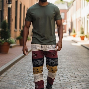 May include: An olive green crew neck t-shirt and patterned trousers in cream, burgundy, and gold. The trousers have geometric designs. White trainers complete the outfit. The person is walking on a cobbled street.