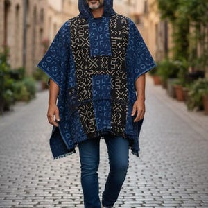 Könnte beinhalten: Ein Kapuzenponcho mit einem Patchwork-Design in Blau- und Schwarztönen. Der Poncho hat einen Fransenbesatz und wird mit blauen Jeans und weißen Turnschuhen getragen. Die Person geht auf einer Kopfsteinpflasterstraße.