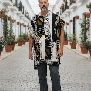 Op de afbeelding: Een man draagt een zwart, wit en goudkleurig patroon poncho met een capuchon en een voorzak. Hij draagt ook een grijze broek en bruine sandalen. De poncho heeft een geometrisch ontwerp.
