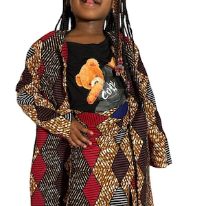 May include: A child wearing a red, black and gold patterned dress with a matching jacket. The dress has a side slit and the jacket has a hood. The child is wearing a black t-shirt with a cartoon bear graphic and the word "Cool" printed on it.