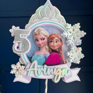 Peut inclure: Un topper de gâteau représentant les personnages Elsa et Anna du film La Reine des Neiges. Le topper est décoré d'une couronne en paillettes argentées, de flocons de neige et du chiffre 5. Le nom "Ariya" est écrit sur une bannière sous les personnages.