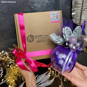 May include: A purple glass Christmas ornament decorated with white faux snow, purple and silver ribbon, and purple and silver beads. The ornament is being held by a person's hands. The ornament is next to a brown box with a pink ribbon and a QR code.