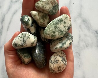 May include: A collection of green and white moss agate tumbled stones. The stones are smooth and polished, with a variety of patterns and colors.