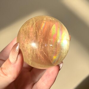 Op de afbeelding: Een doorschijnende, goudgele kristallen bol met interne strepen en iriserende regenboogflitsen. De gepolijste bol wordt in een hand gehouden, wat de natuurlijke schoonheid benadrukt. Een decoratief item.