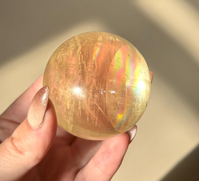 Op de afbeelding: Een doorschijnende, goudgele kristallen bol met interne strepen en iriserende regenboogflitsen. De gepolijste bol wordt in een hand gehouden, wat de natuurlijke schoonheid benadrukt. Een decoratief item.