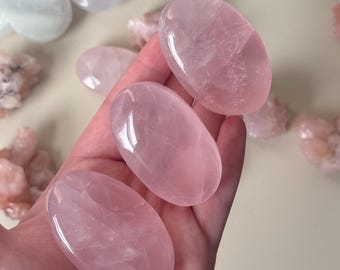 Può includere: Tre pietre di quarzo rosa lucido, di forma ovale e in diverse tonalità di rosa, sono tenute in una mano. Le pietre hanno una superficie liscia e lucida. Altri pezzi di quarzo rosa e pietre a forma di cuore sono visibili sullo sfondo.