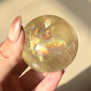 Op de afbeelding: Een doorschijnende, goudgele kristallen bol met iriserende regenboogreflecties. Het gepolijste oppervlak van de bol reflecteert het licht. De kristallen bol wordt in een hand gehouden. Een decoratief item.
